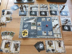 A setup of 'Dead of Winter' with the board centred. The board includes a count of the game's rounds, a floorplan of the colony with a red dice in the middle, and miniature character cut-outs. Around the board are various character and item cards.