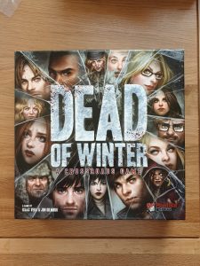 The front of the game's box, with 'Dead of Winter' centred in large silvery letters and flecked with spatters of red. In a circle around the edges of the box are 15 characters, divided by lines of broken ice. 
