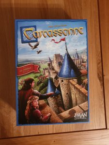 Photo of the game box, featuring a depiction of two characters in medieval dress looking out at the pointed blue turrets of a castle. In the background rises the many different towers of the keep, with green fields and blue sky beyond. The words 'Carcassonne' sit at the top of the box in golden letters. 