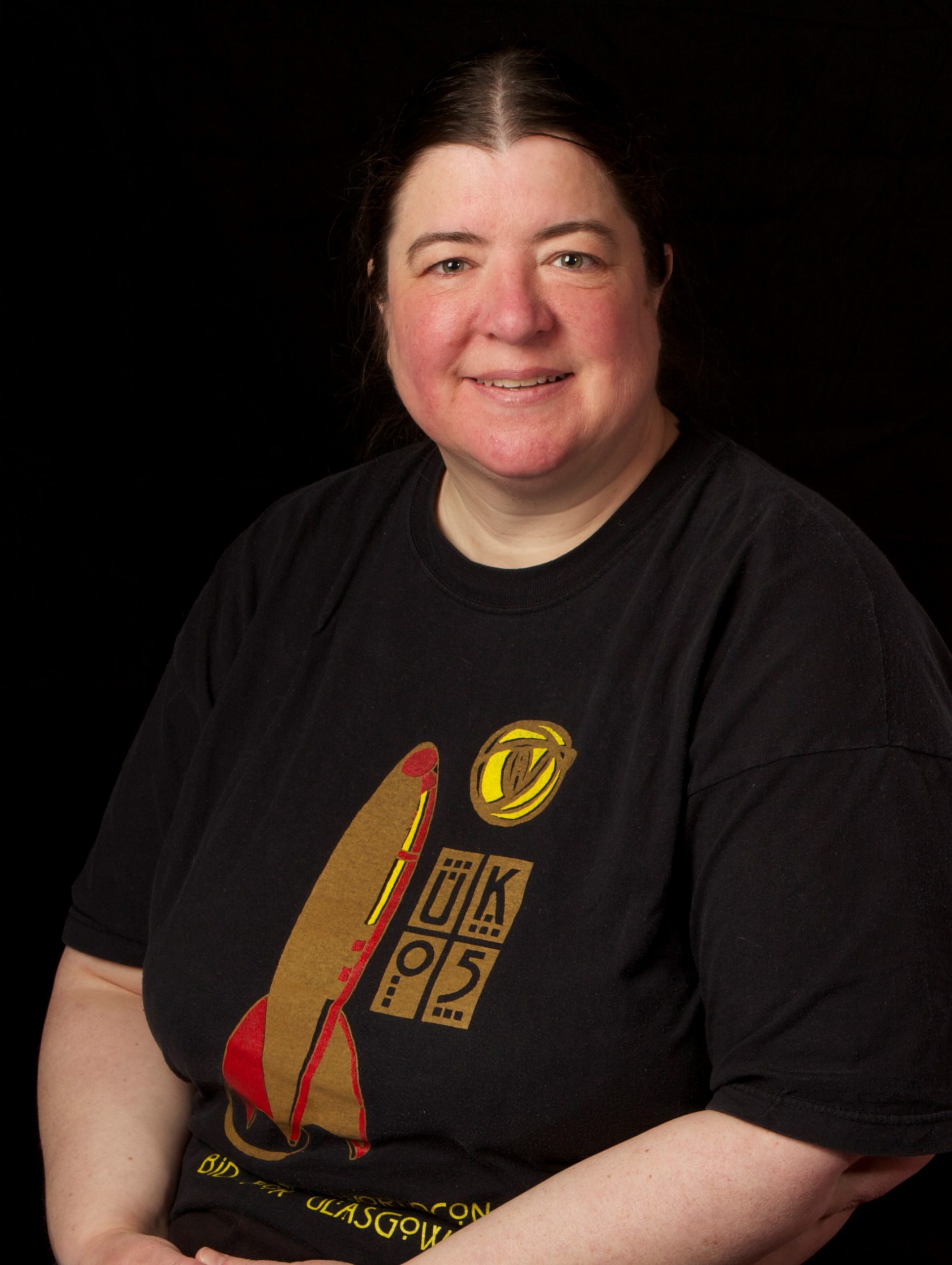 photo of a white woman with rosy cheeks and dark hair in a black t-shirt with a rocket logo on it, she is smiling
