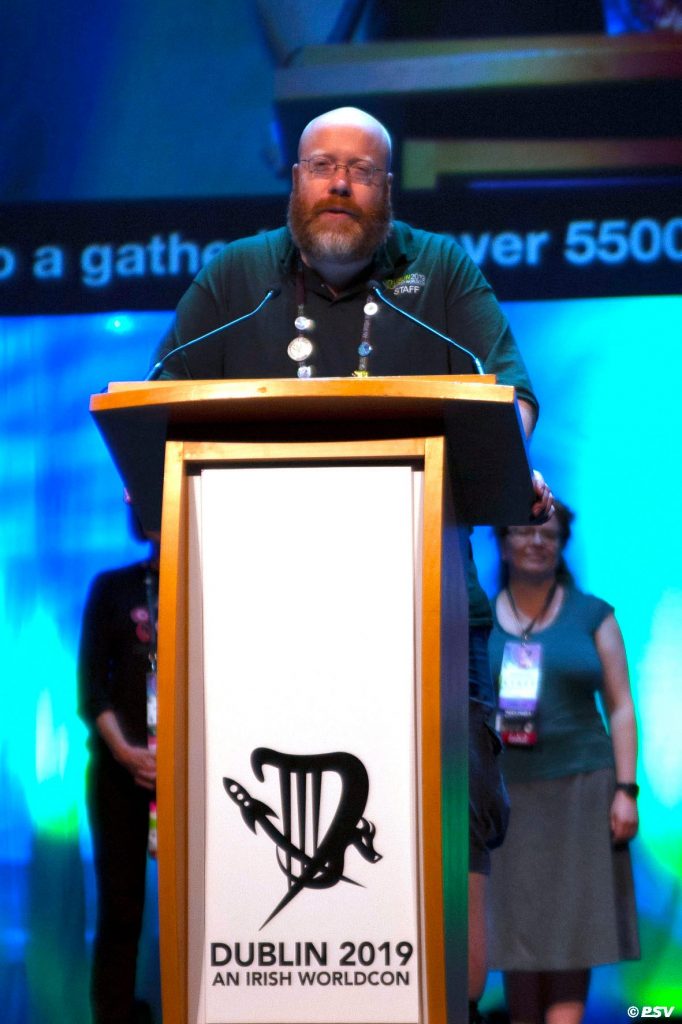 Brian Nisbet standing in front of a lectern with an inscription that reads 'Dublin 2019 An Irish Worldcon'