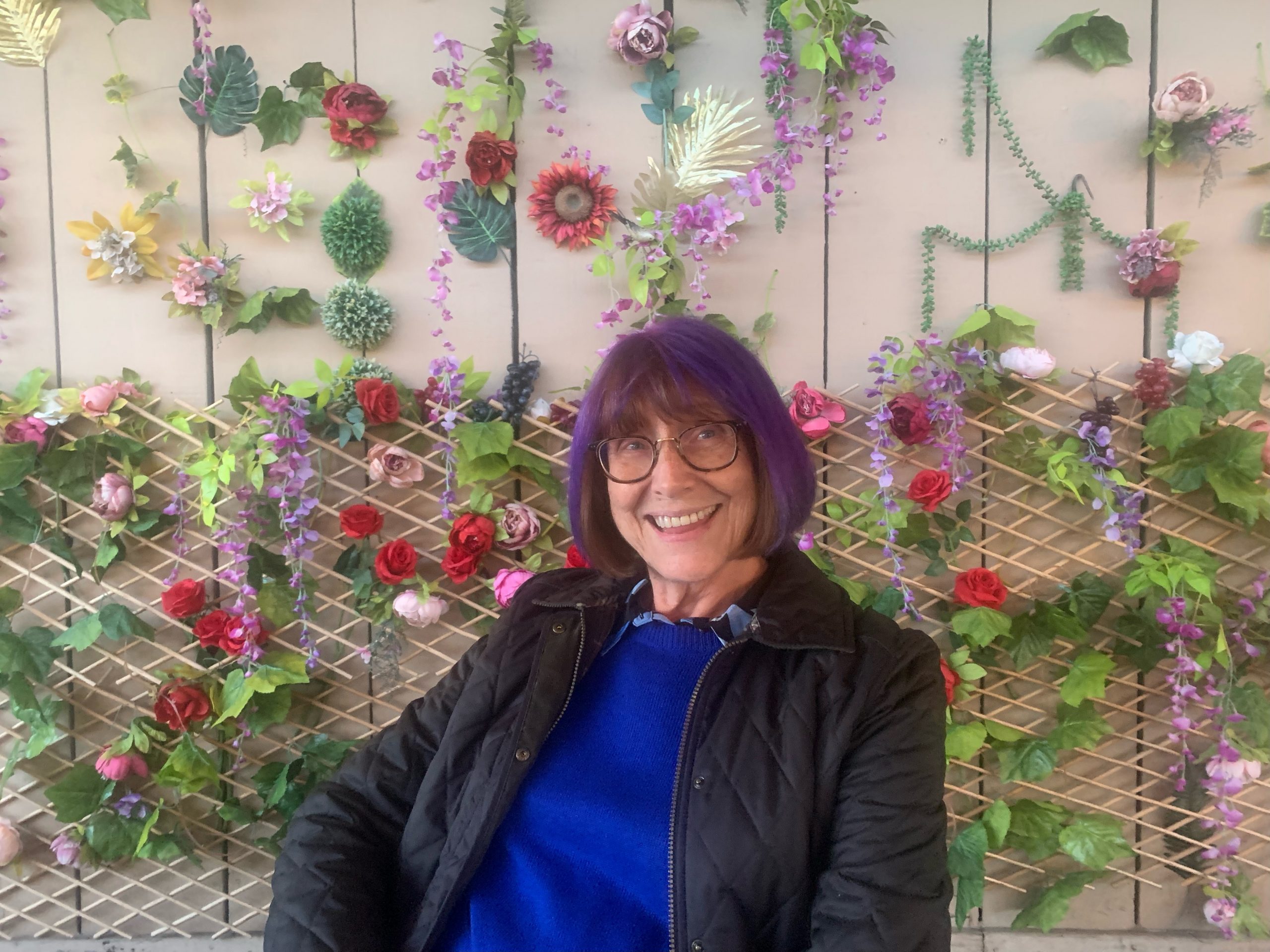 woman in blue top and jacket, with purple bobbed hairstyle, sitting in front of a colourful flower wall. she is looking at the camera and smiling