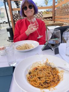 image of woman in red top and with a purple bobbed haircut sitting at a dinner table with a glass of wine, she is wearing large sunglasses, she is looking at the camera and smiling