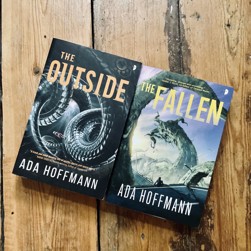 Two books on a wood background, the left features curled up snakes and yellow lettering spelling THE OUTSIDE, the right a snake-like monster from afar and yellow lettering spelling THE FALLEN