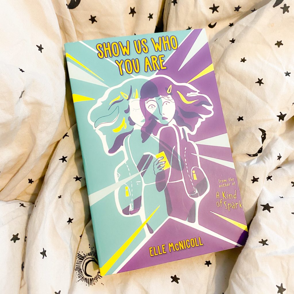 A bright book featuring two silhouettes of a girl in turquoise and purple laid over each other and the title SHOW US WHO YOU ARE in bright yellow, on a light fabric background