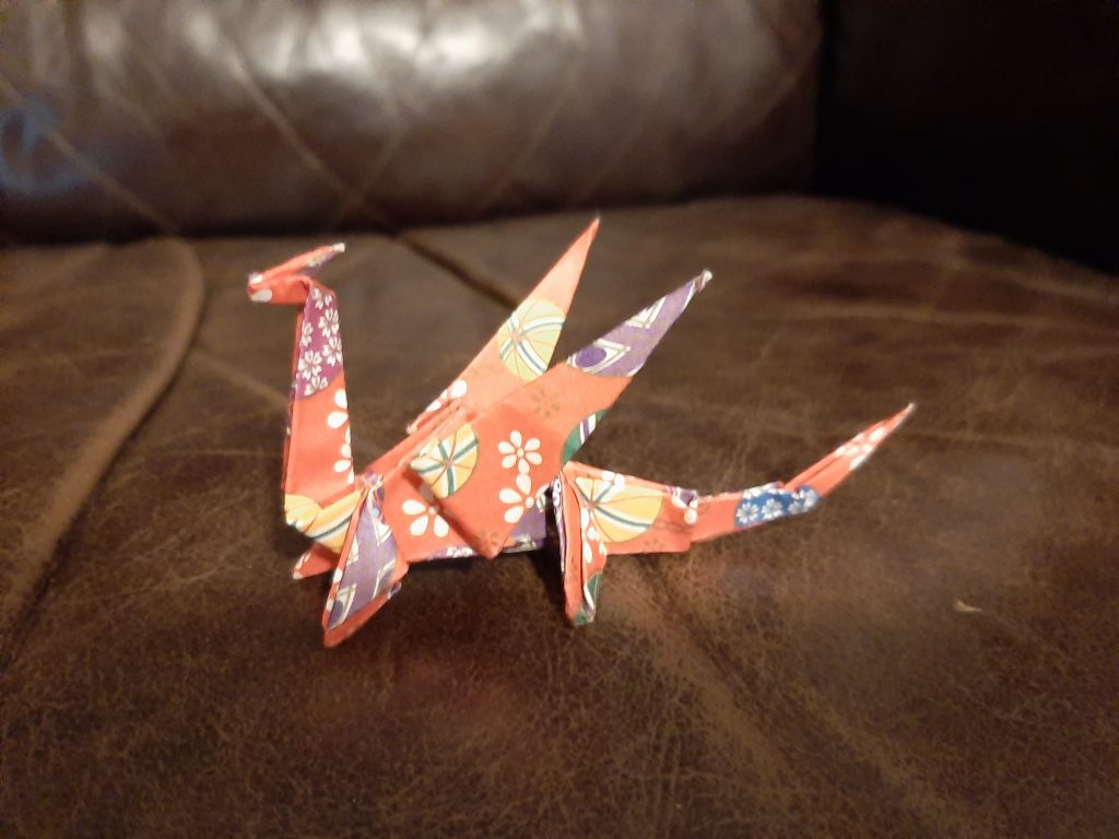 Image shows an origami dragon made out of orange paper with colourful patterns on it in yellow, white and purple