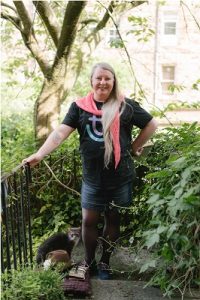 Image shows: Elaine standing in the centre of greenery, her hand is resting on an iron rail. She is wearing a dark tshirt, a denim skirt, and a bright pink scarf. Her long hair is over her right shoulder and she is smiling. 