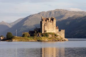 photograph of a castle on a small island on a lake against a mountainous backdrop