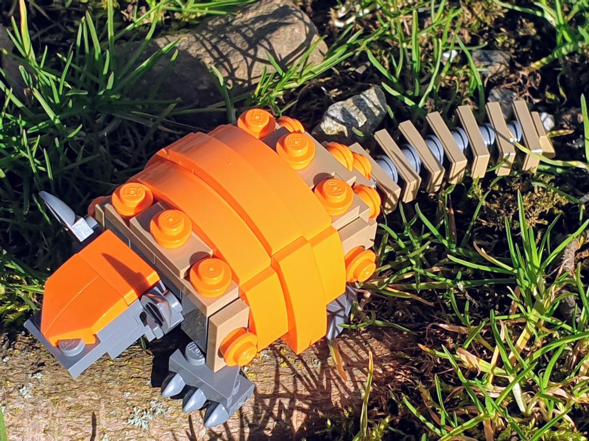 A lego armadillo sitting in the grass. The body is grey and light brown, the armour a bright orange.