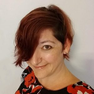 The image shows a head and shoulders photo of Emma K Leadley. She has a pixie cut red brown hair that sweeps over her left eye. She is wearing a black top with large red, grey and white flowers, on a black background. She is smiling.