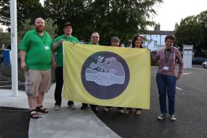 A group of people holding up a yellow flag with a grey circle in the middle. In the centre of the circle there is an image of two clasped hands under the map of Europe.