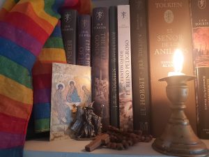 The image shows the close up of a bookshelf. On the left hand side there is a rainbow striped flag draped over some of the books. Next to the flag is postcard with a gilt backgrounded, in the foreground 3 figures sit in robes heads slightly bowed each figure has aureola surrounding their heads. It looks to be a replica of a medieval piece of religious art. Next to that is a a small pertwer statue of a wizard in long robes, a pointed hat, one hand holding a staff, the other is outstretched. The next item on the shelf is a set of wooden rosary beads piled on top of themselves with the simple cross facing outwards. lastly at the far right of the image is a low burning white candle in a pottery holder. There is wax that has spilled on the bottom of the candle holder. The candle is casting a soft light onto the books in the background. These bookes are not all visible but they are different editions of J.R.R Tolkien's work translated into Spanish. Of the books that can be identified there is a copy of The Hobbit and The Silmarillion. 