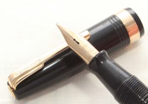 Close up of the very top of a black fountain pen with a golden nib resting on top of its cap. The cap is black with a golden band at the bottom and a golden clip at the top. 