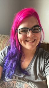 Photo of Esther, a smiling woman with pink and purple hair, wearing glasses and a grey top