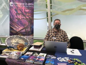 photo of man in chequered shirt and dark facemask sitting at a convention table with promotional material on it