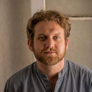 The image shows a head and shoulders picture of Joseph Michael Brennan. He is wearing a dusky bye round collard shirt. His pale golden red hair is short, his beard of the same colour is closely trimmed. He is looking directly forward and is smiling lightly.