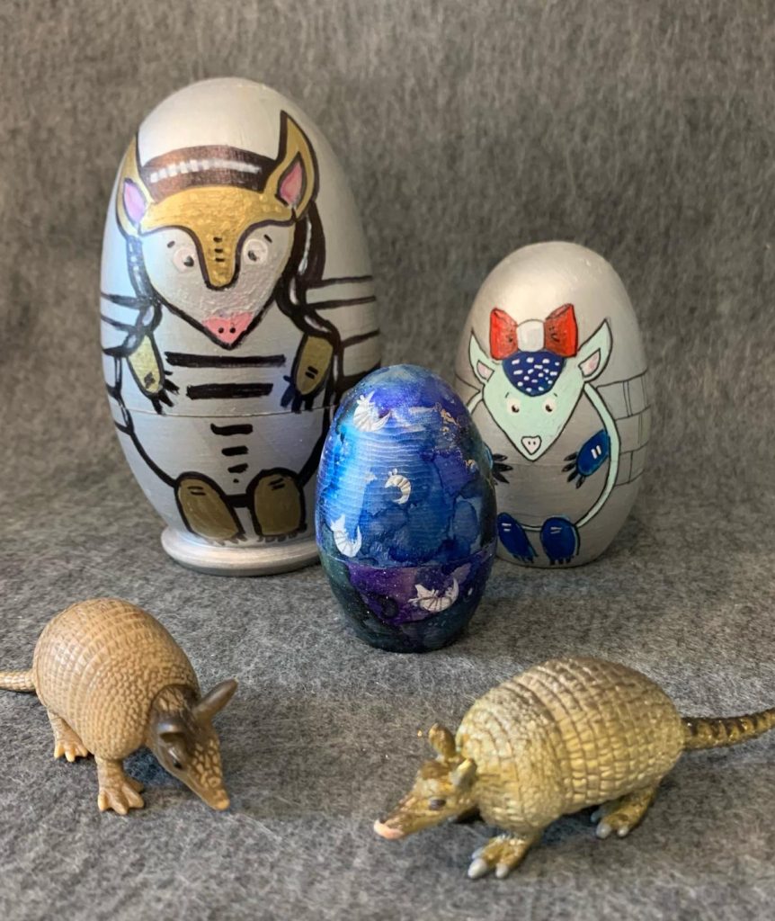 Set of three nesting dolls, painted with armadillos. The biggest on the left in silver with gold details, the middle size on the right with a silver background, light teal head and blue arms and legs. The head is crowned by a big red and white bow. In the middle in front is the smallest doll with many small white armadillos floating on a mottled blue background.