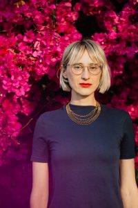 Photo of Marya Brennan. A blonde-haired woman wearing large glasses and a navy top. Behind her is a background of pink flowers.