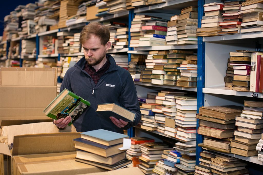 photo of white man in blue swerater sorting books, there is a full case of books behind him