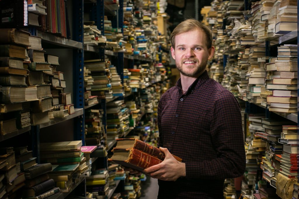 photo of a white man in a plum sweater holding a stack of books, standing between two shelves full of books