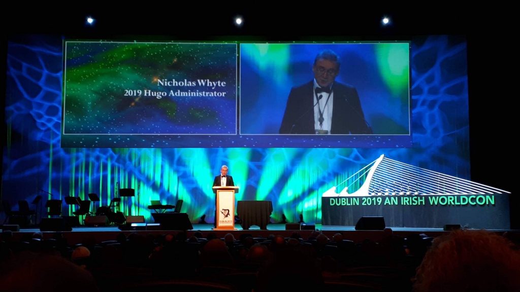 Nicholas Whyte standing at a lectern in a green-lit auditorium. To his right is a sign that reads "Dublin 2019 An Irish Worldcon," and above him his image is projected onto a screen, along with the words "Nicholas Whyte, 2019 Hugo Administrator."