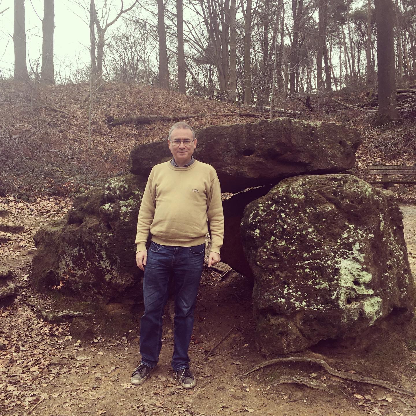 Nicholas Whyte standing in a woodland area. Immediately behind him are some large boulders, and further behind is a hillside with trees and fallen leaves.