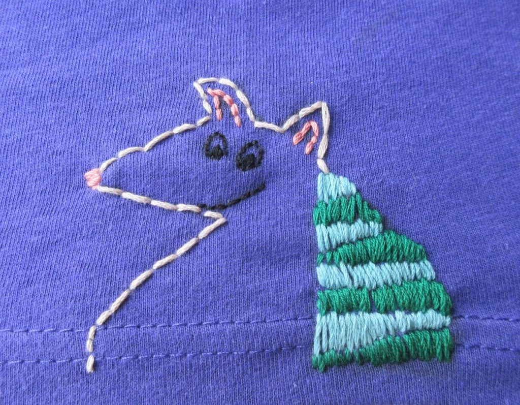 Stylized armadillo embroidered on the seam line of a purple Glasgow in 2024 tshirt. The body is outlined in cream, and the armour has alternating green and light blue stripes.