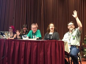 An image taken at a panel at a con. Five people sat at a table, waving at the camera and looking very excited to be there