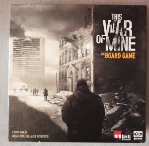 The front of 'This War of Mine' box, with the title in large grey letters at the top right. The image of a war-torn cityscape covers most of the box, with a shadowed figure in the foreground, looking towards rubble, a dilapidated building, and - in the background - a smoking skyscraper. 