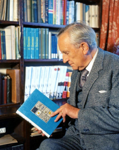 JRR Tolkien holding a blue book in front of book cases. Image credit The Tolkien Society