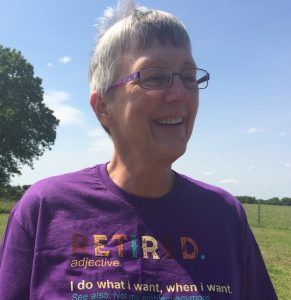 The image shows a head and shoulders shot of Robin Reid. She has cropped short grey hair that is being blown up by a breeze and is smiling. She is wearing purple framed regtangular glasses and a purple tshirt. On the Tshirt there is the following text in luticoloured lettering "RETIRED. adjective. I do what i wasnt, when I want." The remainder of the text is cut off. Behind her on the left is the edge of a large tree, a bright blue sky and green feilds. 