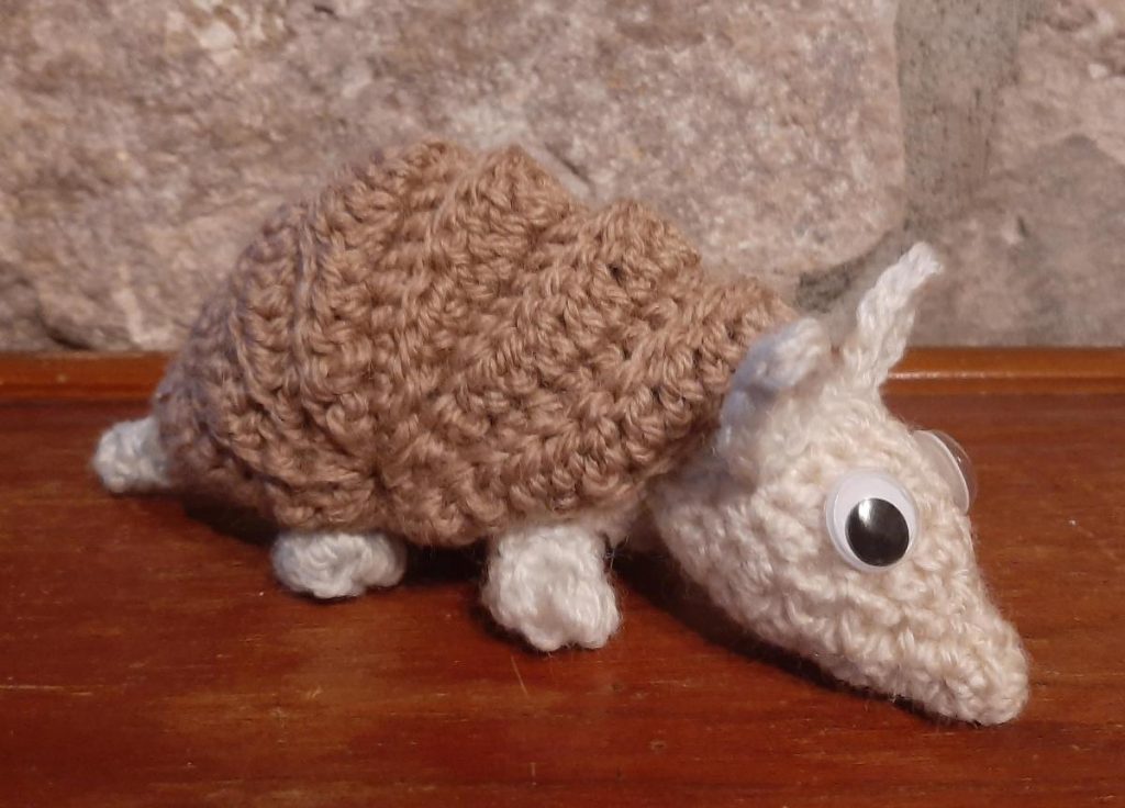 Tiny crochet armadillo with a cream body, light brown armour and big googly eyes.