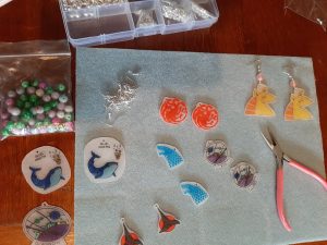 Work space with the finished pendants, earrings and jewellery supplies: different types of beads, earring hooks, and round-nose pliers.