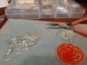 Close-up of a Game of Thrones pendant, the earring hooks and split rings, and the pliers. Box of additional jewellery supplies in the background.