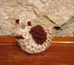 A small crochet snail with a beige body and a brown shell sitting on a wooden table.