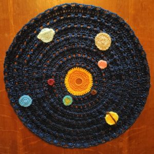 Round crochet of solar system: orange and yellow sun in the middle, dark navy sparkly background, and all the planets attached in different positions around the sun.
