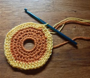 Crocheted sun with orange center worked around keyring and one round of yellow added. Blue crochet hook in open loop.