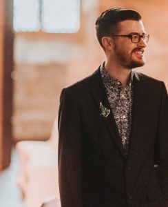 The images shows an upperbody porttrait shot of Taylor Driggers. He is wearing a charcoal grey suit, a grey and white patterned shirt. on the lapel of the suit is a the leaf pin given to hobbits in Lord of the Rings. He is looking to the right and smiling to something off camera. He is wearing rectangular glasses, and his brown beard is short and tidy. His brown hair is shorter on the sides, longer on the top and swept from right to left.