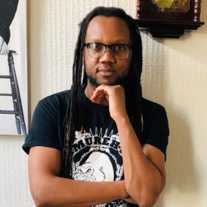 Portrait of a Black man with long dreadlocks and glasses, wearing a black t-shirt in front of a wall