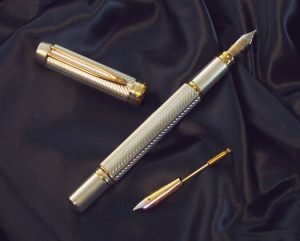 Image of the parts of a silver and bronze fountain pen sat on a navy-coloured cloth. In the middle is the pen itself, to its left is the cap, and to its lower right is a secondary bronze nib.