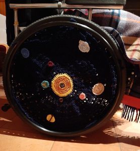 A wheelchair wheel with a dark blue crochet cover attached, showing the sun in the middle and the planets of the solar system around it.