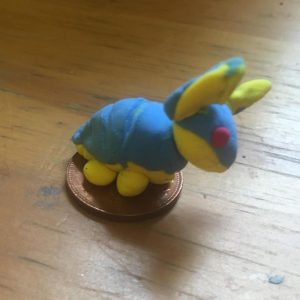 A tiny polymer clay armadillo with a yellow body and blue armor sitting on a penny coin on a wood background.