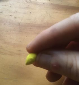 Smaller teardrop-shaped ball of yellow clay, presented between thumb and index finger of a comparatively large hand.