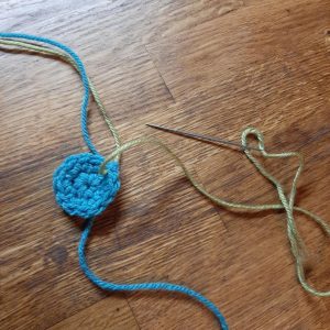 A light blue crochet circle, with a green thread and an embroidery needle attached.