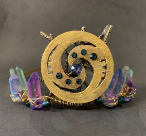 Front on view of a tiara made of crystals - all different tones of purple- with a gold centre piece of CoNZealand's swirling logo bedecked with small blue crystals.