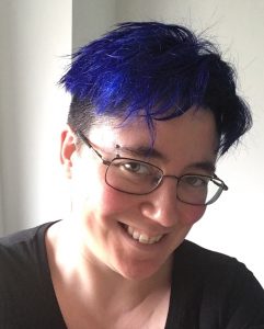 The image is a head and shoulders photo of Juliet Kemp. She is wearing a black t-shirt, and metal rimmed glasses. She has beautiful bright blue hair with a dark undercut, there is a bar peircing over her left (on image) eyebrow. She is smiling and the left side of her face is bathed in light.