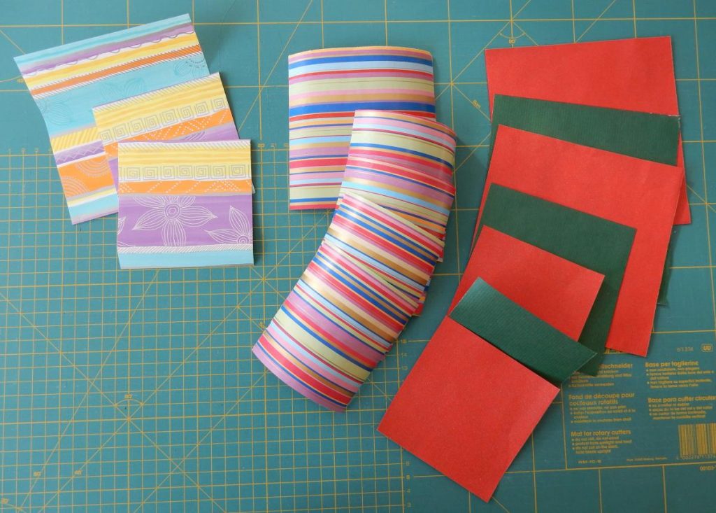 Lying on a green cutting mat is a selection of paper squares made from gift wrapping papers - left a striped pastel with printed graphic patterns, in the middle a multicolour striped paper, on the right a solid with red on one side and green on the other.