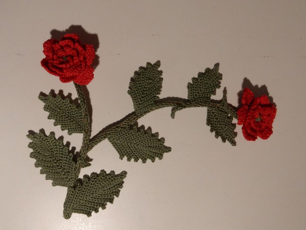 A sprig of crochet roses with serrated leaves and two red flowers at the end of the splitting branches.