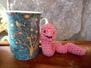 A small pink crochet worm leaning against a mug with a tree of life pattern printed on it.