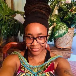 Nnedi is a black woman with large black rimmed glasses and a tattoo on her upper right arm. Her hair is tied up under a black scarf with some dreadlocks wrapped around the outside of the scarf. She is wearing a colourful tank top and is smiling at the camera